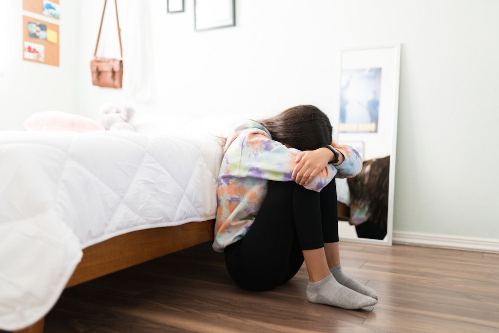 A teen sitting on the floor by her bed during the day, appearing overwhelmed